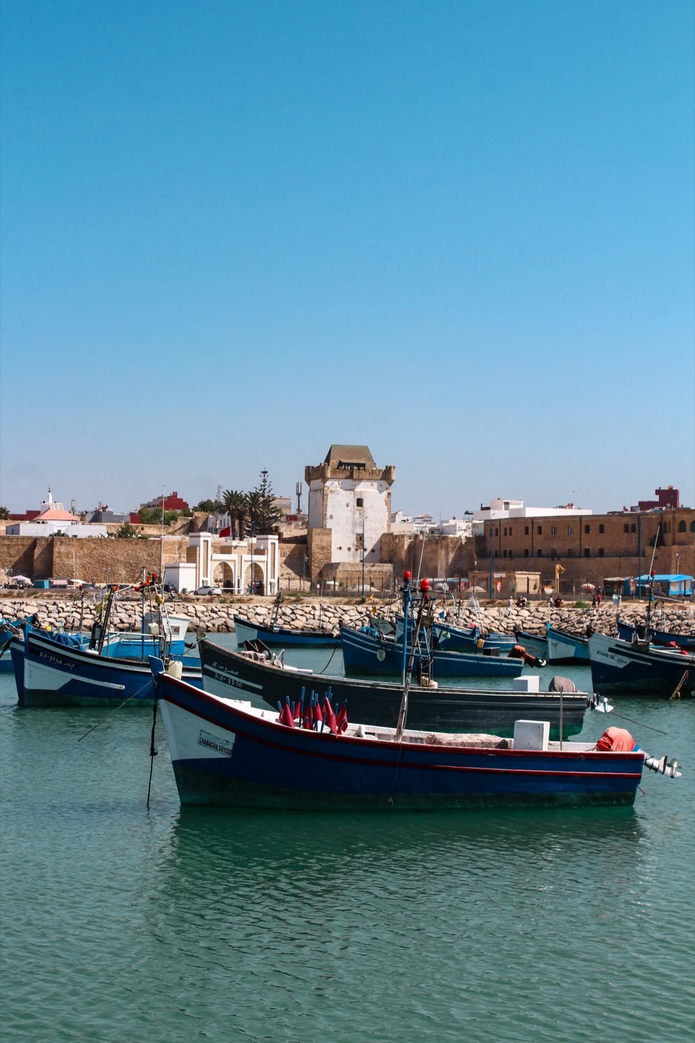 Blue and white fishing boats in the Asilah harbor, with the ramparts and tower of the medina behind them