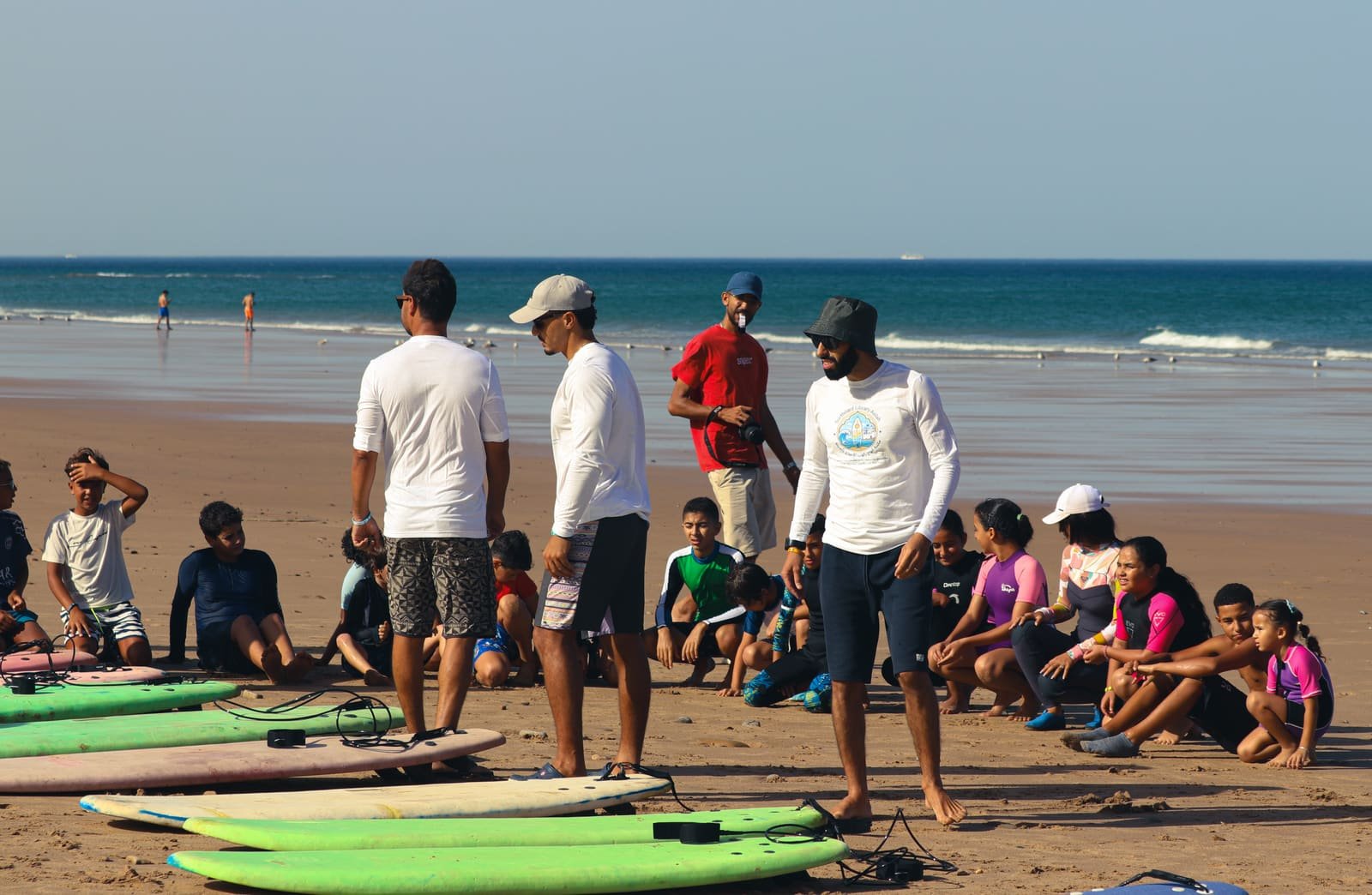 Kids sitting on the sand behind a row of foam surfboards while instructors brief them before a session at Sidi M'Ghait Beach