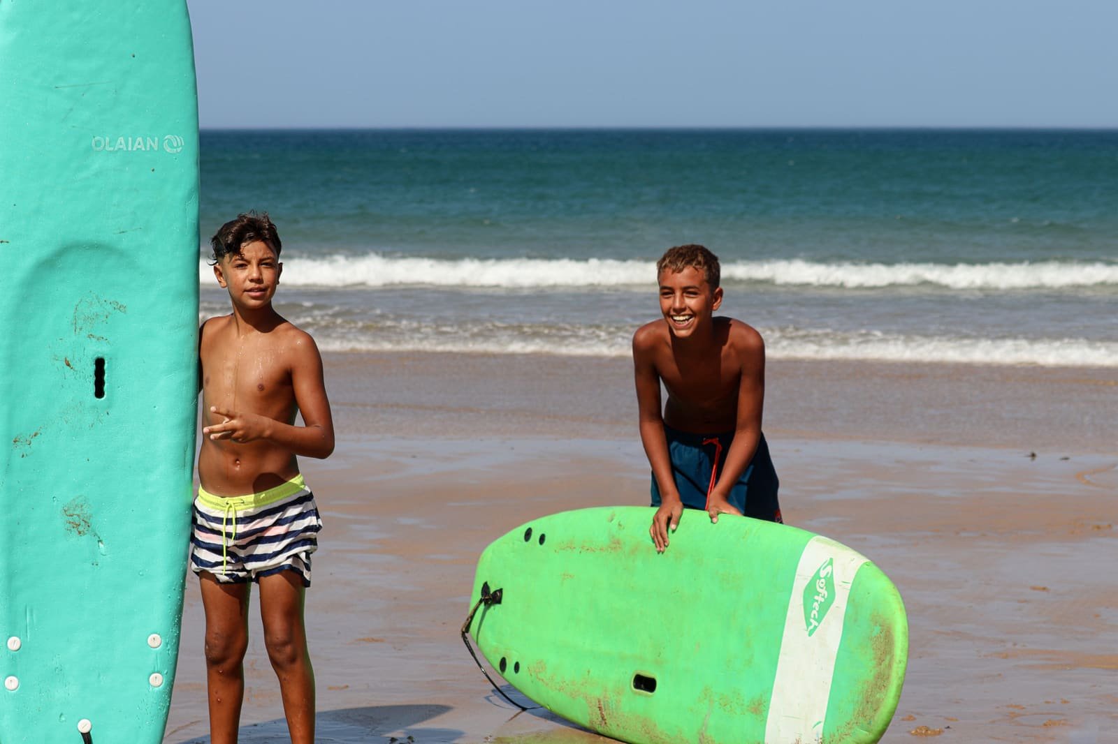Two boys smiling on the sand beside their foam surfboards