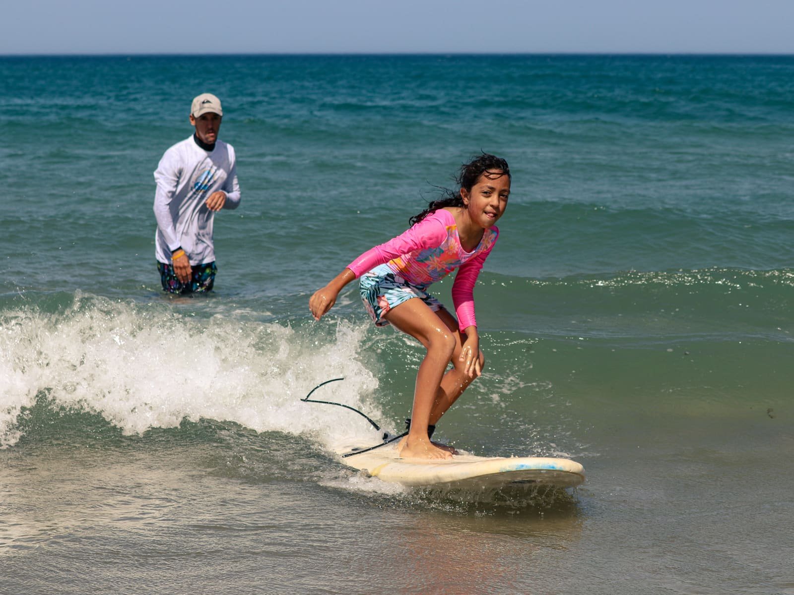 A girl standing up on a surfboard in the whitewater while an instructor watches from behind