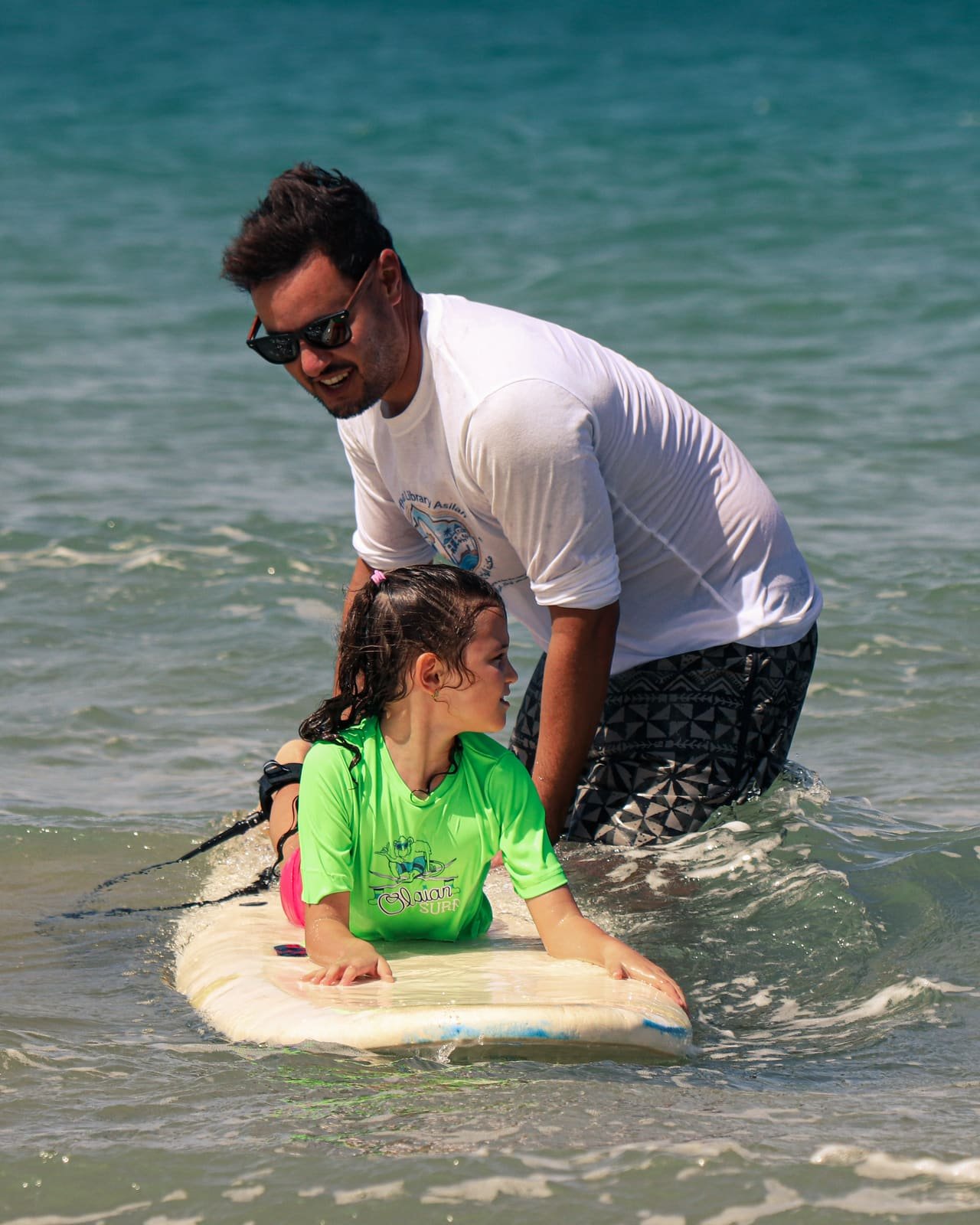 An instructor steadying a young girl on a foam surfboard in shallow water