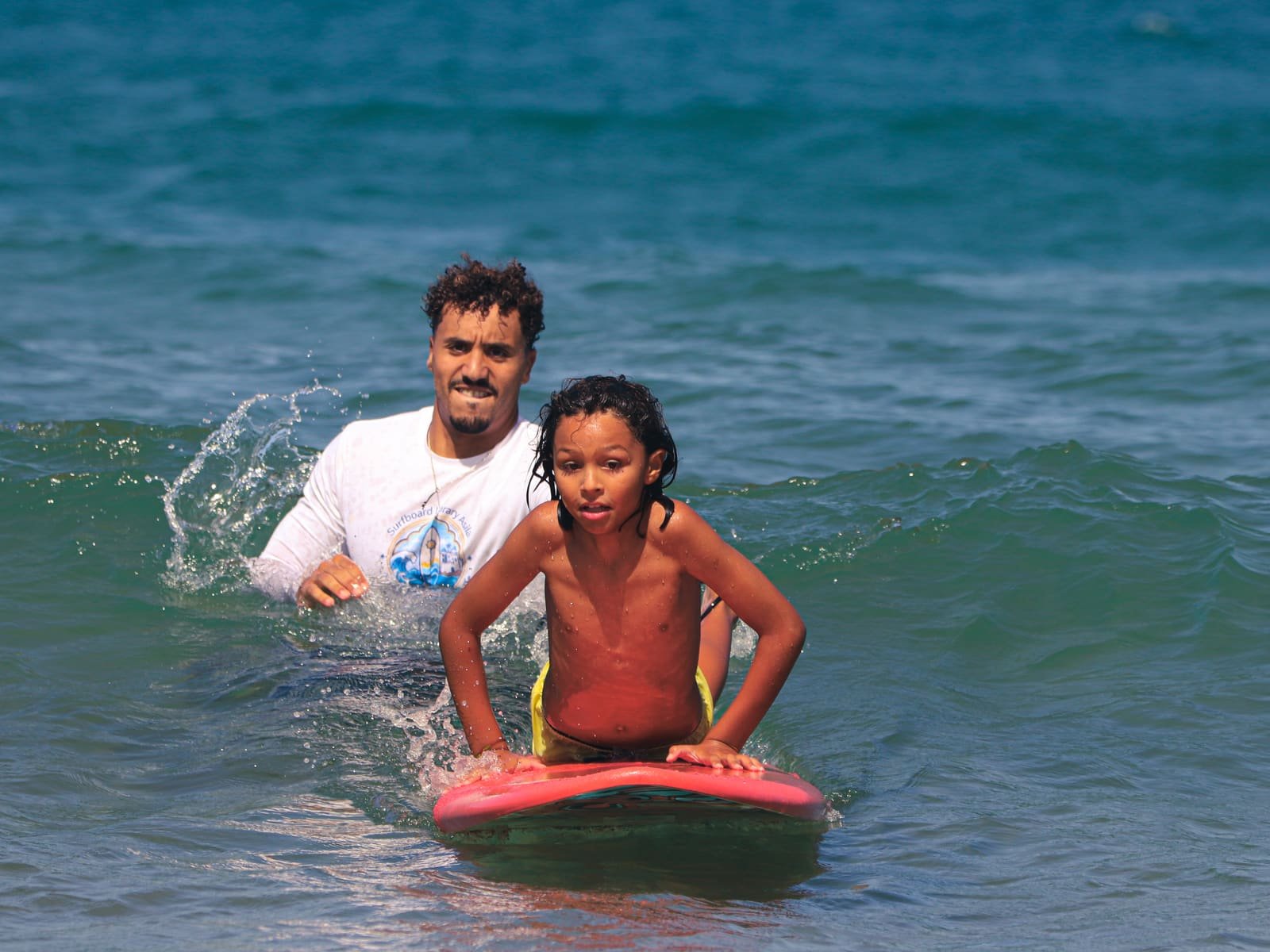 A smiling instructor pushing a kid into a wave on a red foam surfboard