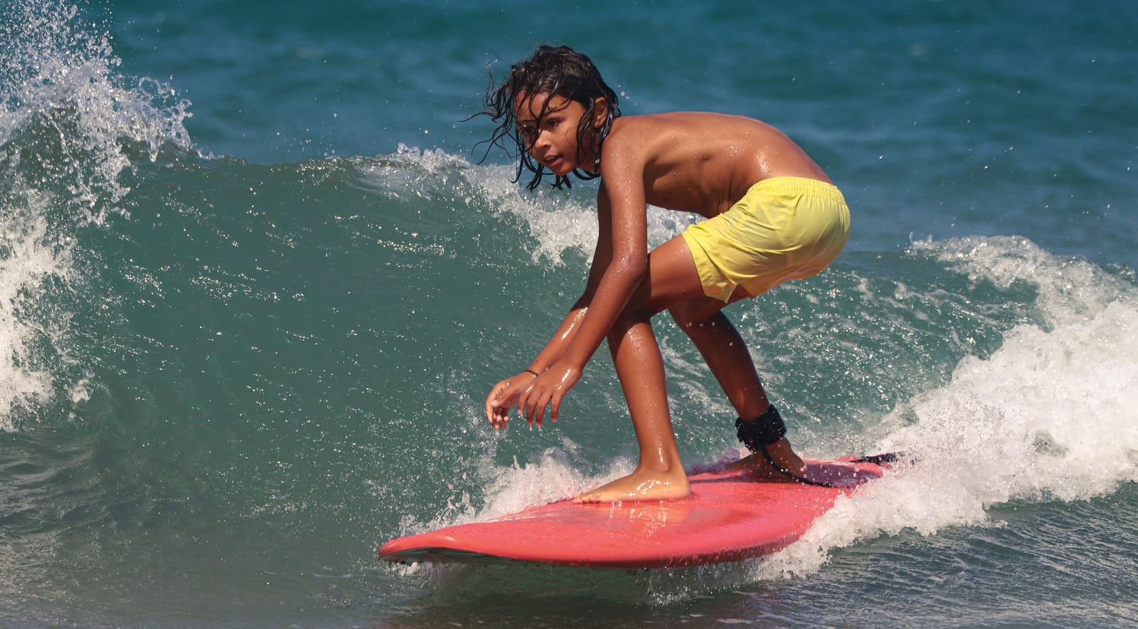 A kid crouched low on a red foam surfboard, riding along a small wave