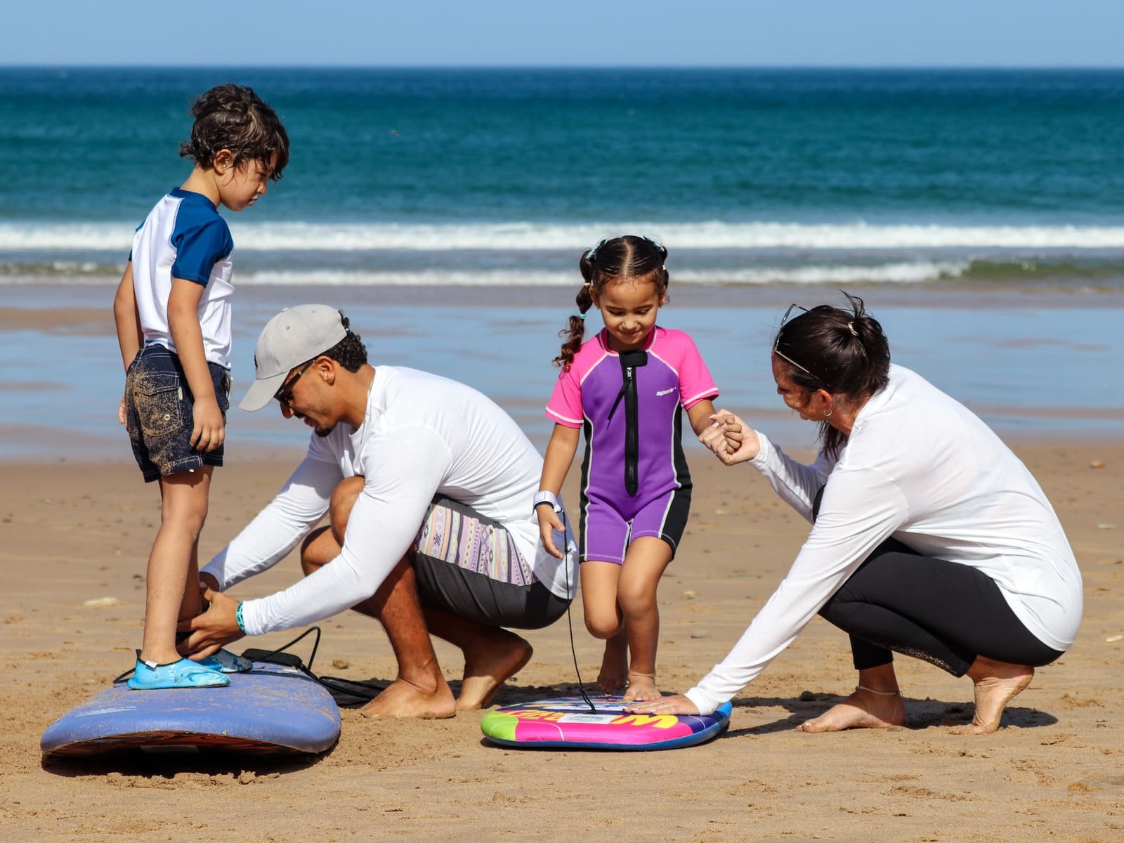 Two instructors kneeling in the sand, showing young kids where to stand on a foam board