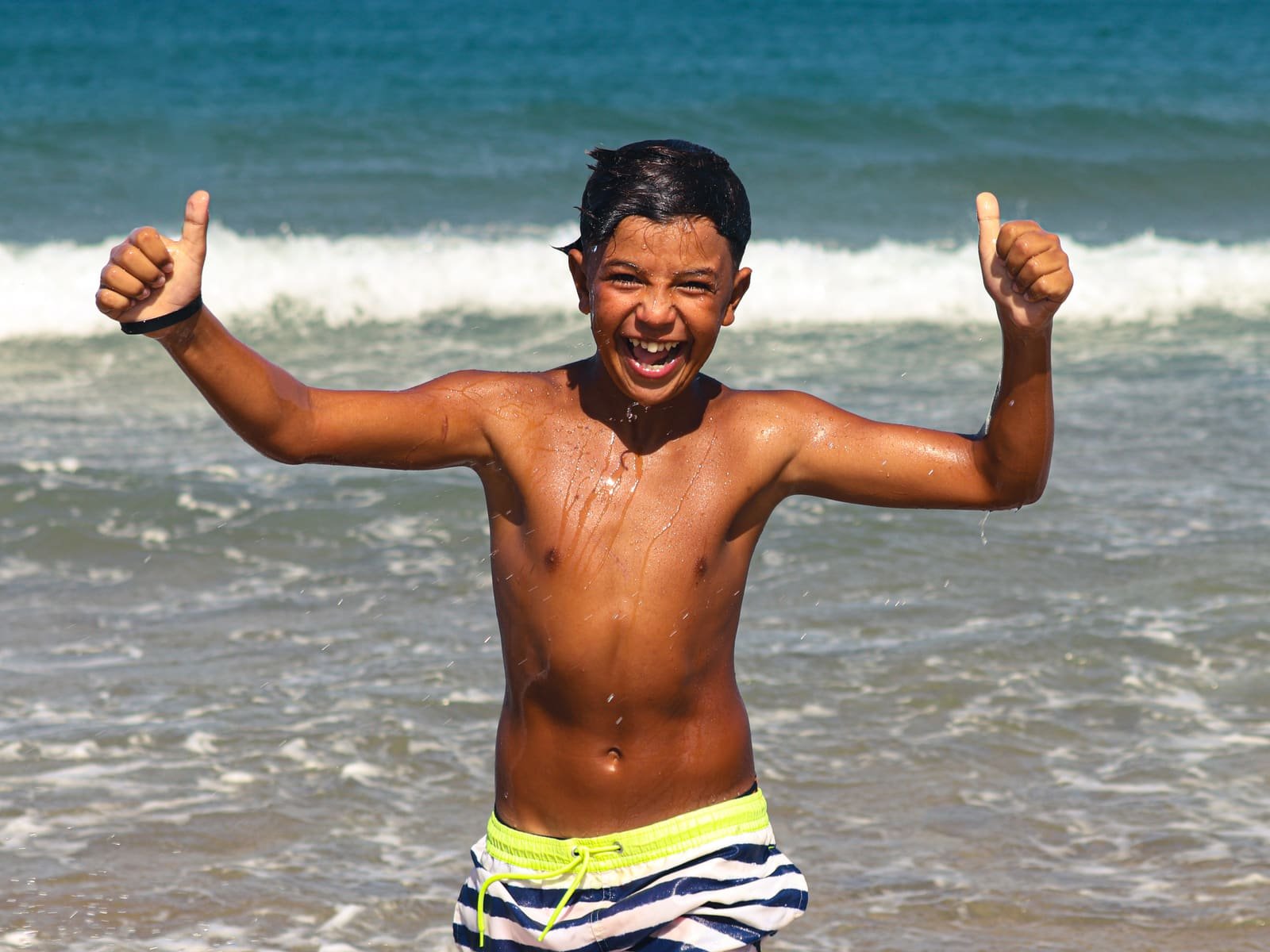 A boy fresh out of the water, grinning with two thumbs up