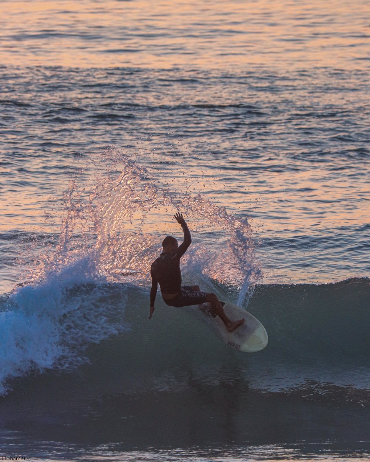 A surfer silhouetted against a pink and gold sunset, turning off the top of a wave