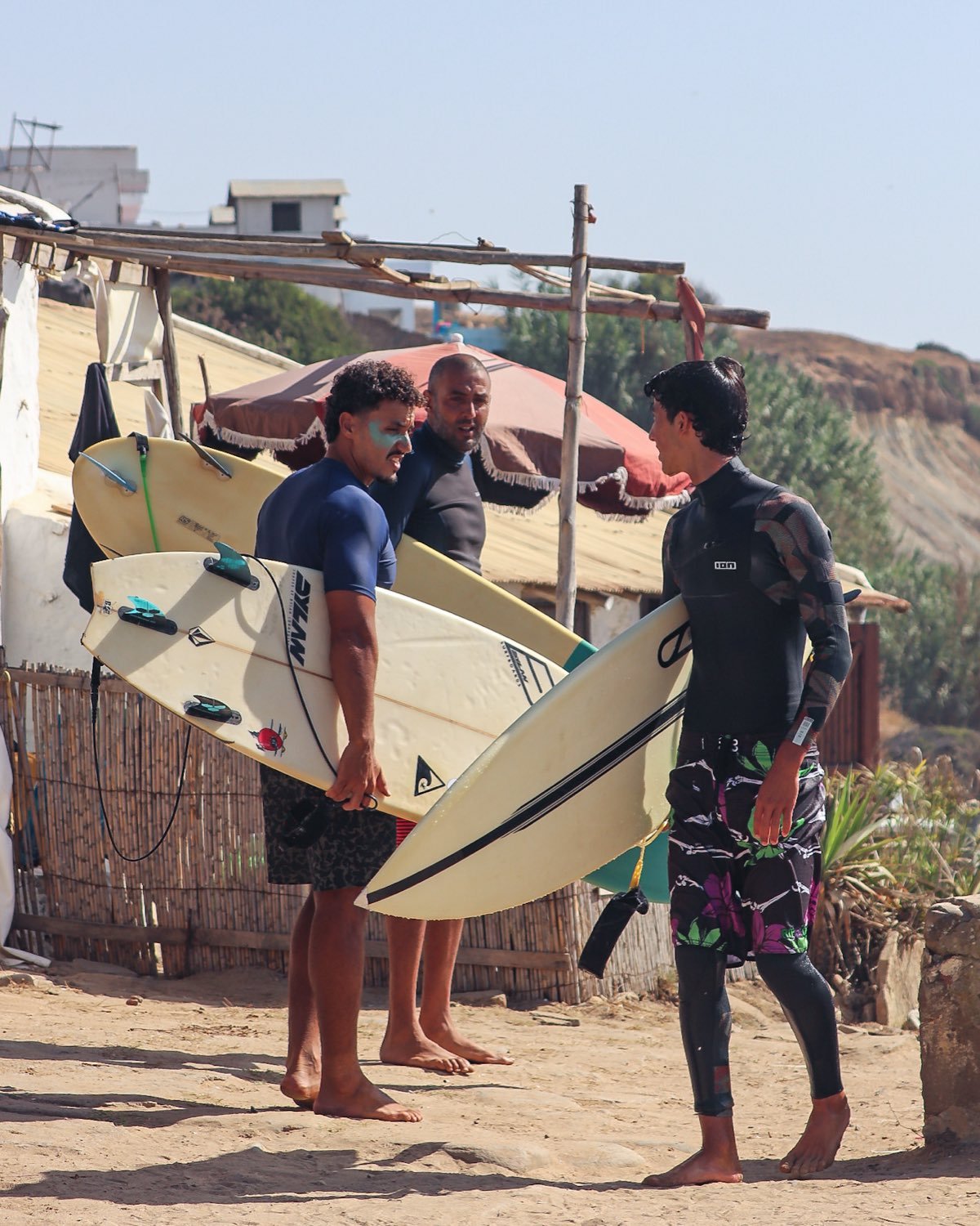 Three surfers carrying their boards up the sand past a beach shack in Morocco